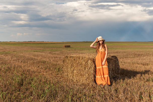 curly young rural teenager girl stands near a bundle of hay in a sundress and hat on a harvested wheat field with a backdrop of a sunset rain sky and a rainbow - Foto, Bild