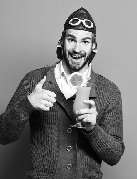 handsome bearded aviator man with long beard on smiling face holding glass of alcoholic cocktail in gray knitted sweater with hat and glasses on red studio background - Фото, зображення
