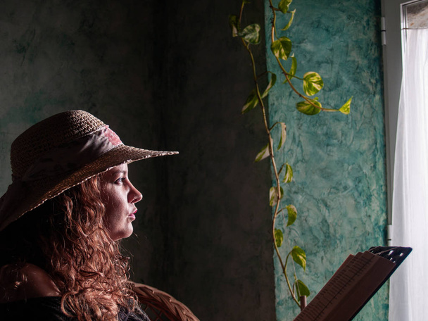 Portrait of a young woman with curly hair wearing a straw hat with natural light coming in through a window in a rural house - Photo, Image