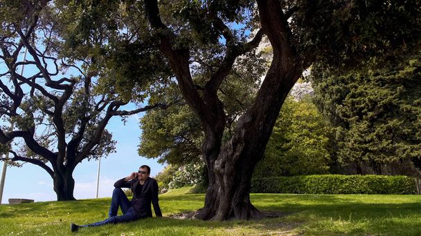 Бизнесмен сидит на траве и разговаривает на смартфоне, отдыхает в парке
 - Фото, изображение