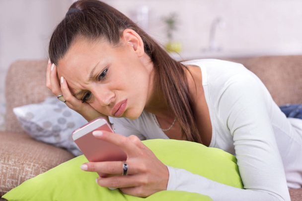 Mujer joven y bonita tumbada en el sofá y mirando el teléfono móvil. Expresión infeliz en la cara
 - Foto, Imagen