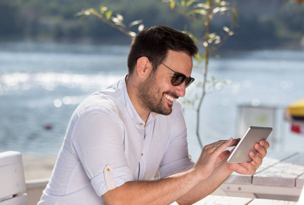 Красивый мужчина сидит в кафе на пляже и смотрит на планшет. Наслаждаясь летним утром у реки
 - Фото, изображение