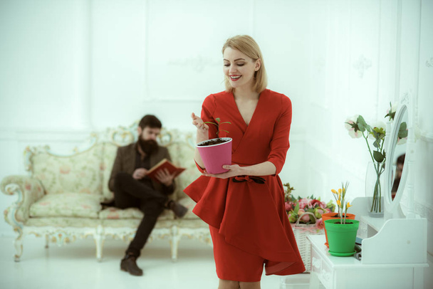 Αυξανόμενη έννοια. Σέξι γυναίκα κρατήστε λουλούδι αυξάνεται σε κατσαρόλα ενώ ο άνθρωπος διαβάζοντας το βιβλίο στον καναπέ. Ποτ καλλιέργεια φυτών. Ενεργού καλλιεργητική περίοδο - Φωτογραφία, εικόνα