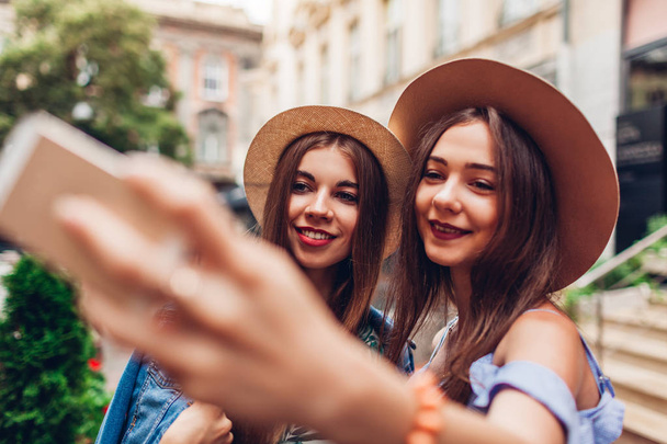 Outdoor portrait of two young beautiful fashionable women taking selfie using phone. Happy girls having fun in city. Best friends wearing hats - Photo, Image