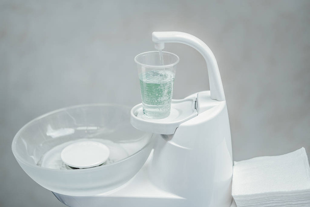 Close up of white dentist spittoon and plastic cup with light green water. - 写真・画像