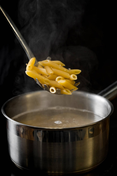 Chef lowers a portion of penne pasta into a saucepan with boiling water close-up on a black background - Фото, изображение