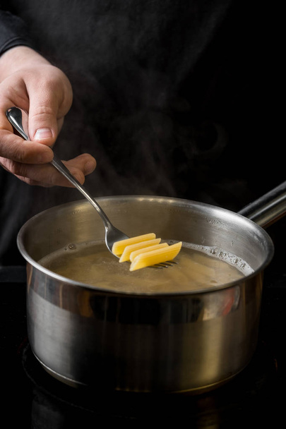 Chef lowers a portion of penne pasta into a saucepan with boiling water close-up on a black background - Fotografie, Obrázek