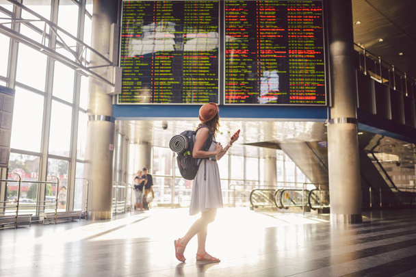 Utazási téma és tranosport. Gyönyörű fiatal kaukázusi nő a ruhát, és állt befelé vasútállomás, illetve a terminál látszó-on egy menetrend gazdaság egy piros telefon, hátizsák, használja a kommunikációs technológia - Fotó, kép