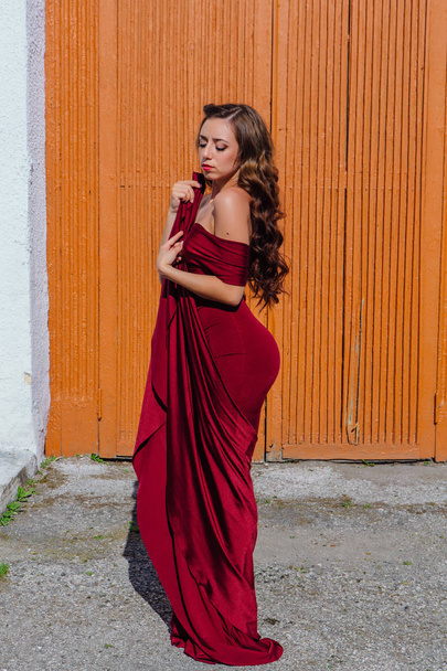 Beautiful young woman in a long red evening gown with a train standing next to the old door - Foto, imagen