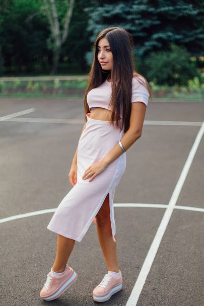 Charming brunette female dressed in a pink dress posing on the basketball court during sunset. - Foto, Bild