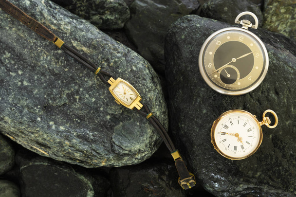 Set of watches on wet green stones with a classic gold pocket watch a black and silver pocket watch and a wristwatch with worn leather straps - Photo, Image