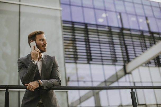 Молодий успішний бізнесмен в сірому костюмі і тримає свій смартфон, стоячи біля сучасного офісу або хмарочосів
 - Фото, зображення