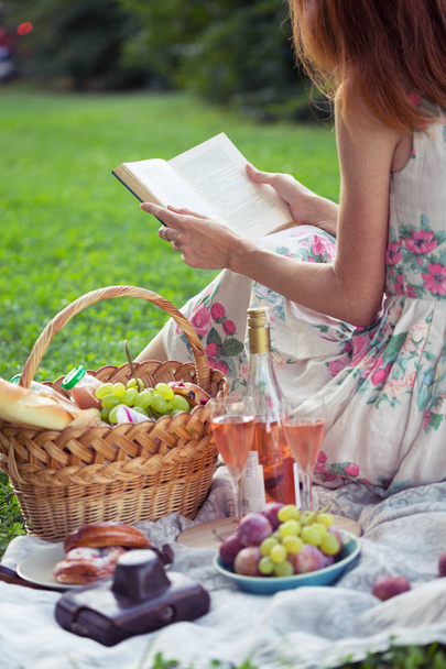 Summer - picnic in the meadow.  girl sitting reading a book and near a picnic basket and baguette, wine, glasses, grapes and roll - Photo, Image