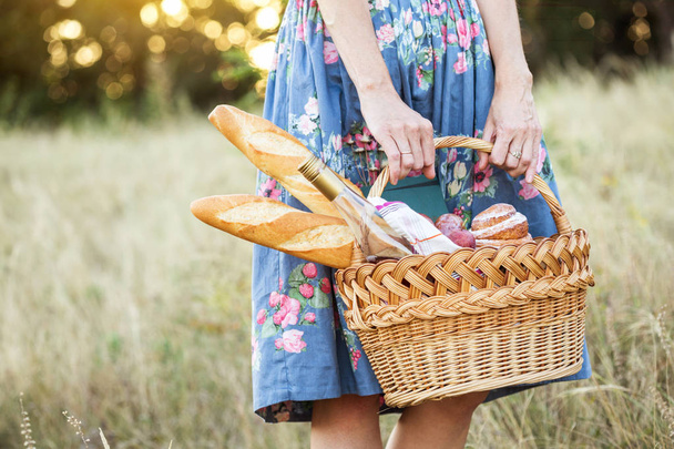 Summer - picnic in the meadow.  girl holding a basket for a picnic with baguette, wine, glasses, grapes and roll - Fotó, kép