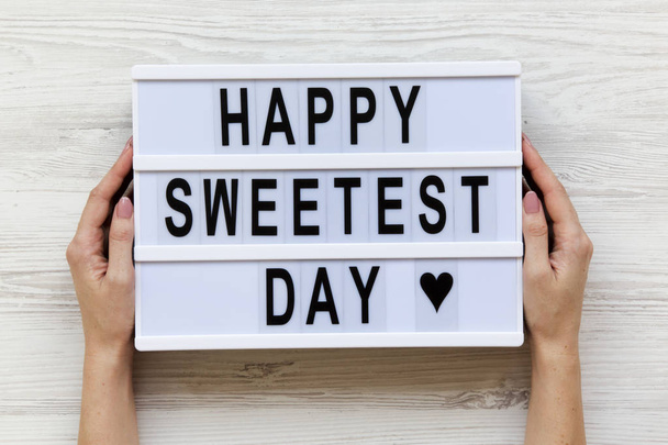 Female hands hold modern board with text 'Happy Sweetest Day' word over white wooden background, top view. From above, flat lay, overhead.  - Photo, Image