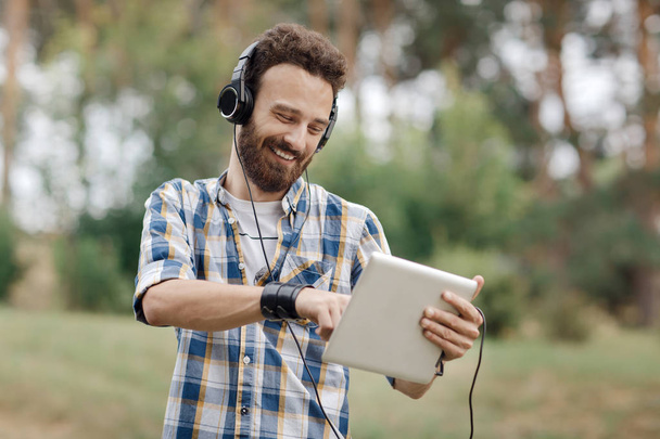 Young bearded hipster with tablet and headphones listening music in the park. - Foto, Imagem