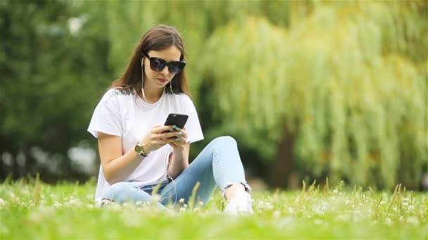 Mujer linda está leyendo el mensaje de texto en el teléfono móvil mientras está sentado en el parque. - Imágenes, Vídeo