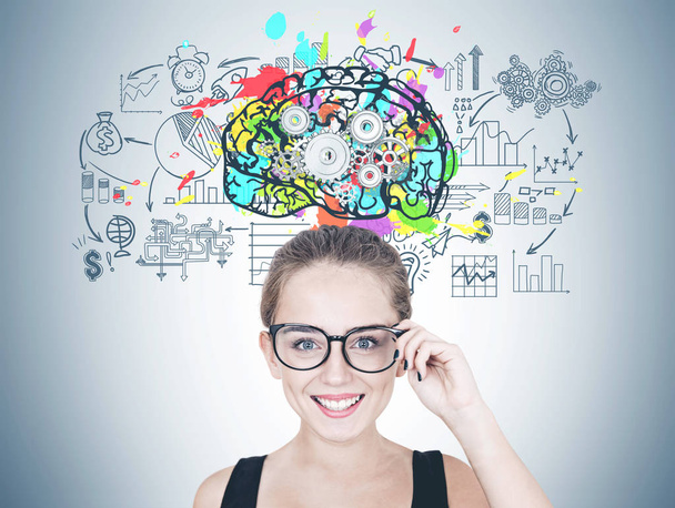 Portrait of a cheerful young woman touching her glasses and looking at camera. Black tank top and long hair in ponytail or bun. Colorful brain with gears and business planning icons on a gray wall. - Photo, Image