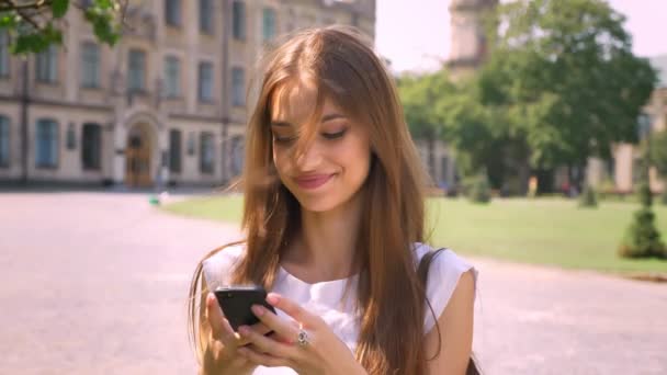 Sexy lady is standing in park in daytime, typing message on smartphone, watching at camera, smiling - Imágenes, Vídeo