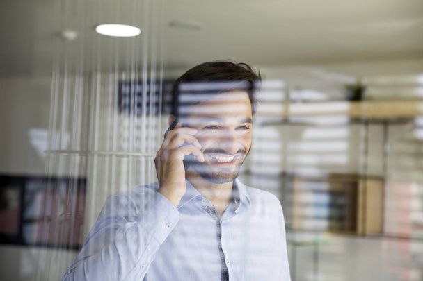 Pohledný mladý muž používající mobilní telefon za sklem v kanceláři - Fotografie, Obrázek