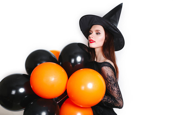Portrait of beautiful sexy young woman in black witch halloween costume with orange and black balloons over white background - Foto, Bild