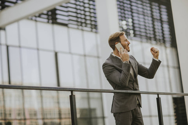 Joven empresario exitoso con traje gris y sosteniendo su teléfono inteligente mientras está de pie cerca de la oficina moderna o rascacielos
 - Foto, imagen