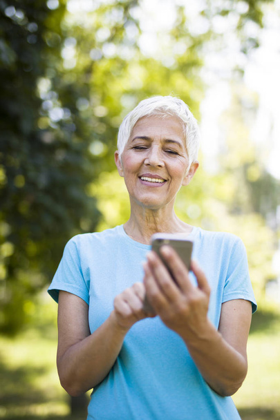 Portret sportowy senior kobiety przy użyciu telefonu komórkowego w parku - Zdjęcie, obraz