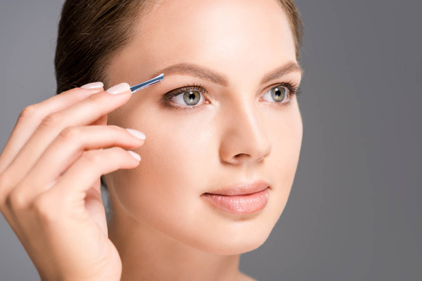 portrait of young woman plucking eyebrows with tweezers isolated on grey - Fotoğraf, Görsel