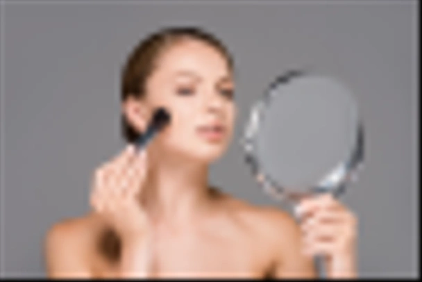 portrait of young woman looking at mirror and applying blush isolated on grey - Fotografie, Obrázek