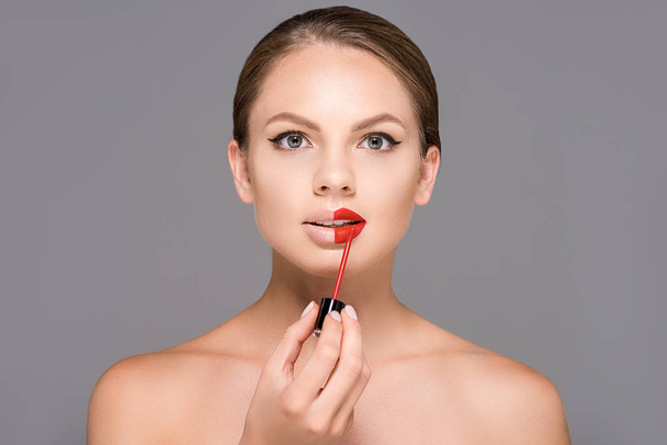 portrait of beautiful young woman with bare shoulders applying red lipstick isolated on grey - Photo, image