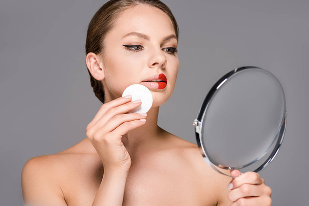 portrait of young woman with red lipstick on half of lips and sponge looking at mirror isolated on grey - Photo, Image