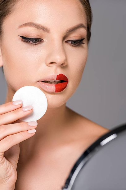 portrait of young woman with red lipstick on half of lips and sponge looking at mirror isolated on grey - Foto, imagen