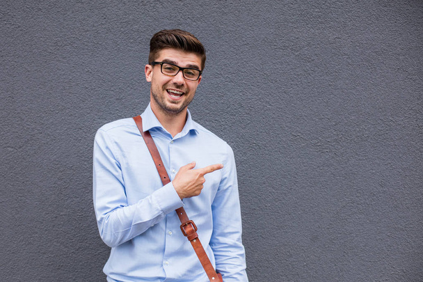 pointing the empty side. handsome smart casual man with eyeglasses and leather bag outside pointing a free space next to him with confidence and smile - Foto, imagen