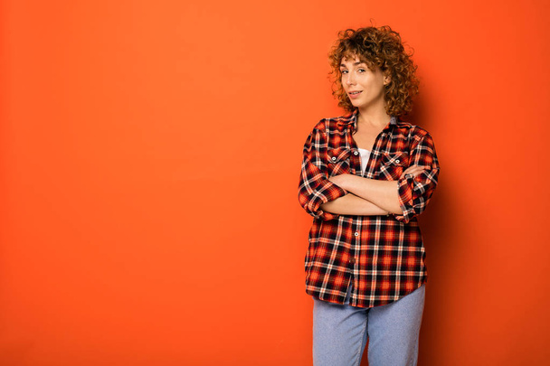 pretty curly woman in a checkered shirt and jeans standing over an orange background with empty space for text next to her with folded arms - Фото, изображение