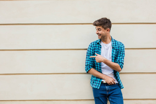 handsome man in checkered shirt smiling standing outside leaning on a wall with free space next to him pointing the empty area - Fotoğraf, Görsel