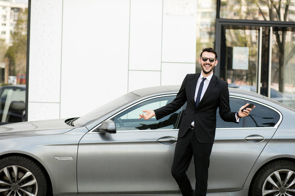 uber driver in elegant suit in front of an luxury car showing his cellphone, outside on the street in front of an office building - 写真・画像