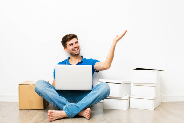 young man in blue shirt sitting on the floor next to his cardbox, looking on internet about apartments, mooving to new house. on white background - Фото, зображення