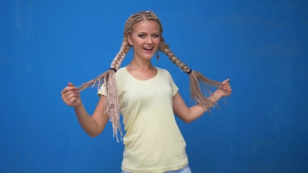 Trendy hipster girl shaking her braided hair pigtails and spinning around on blue background - 映像、動画