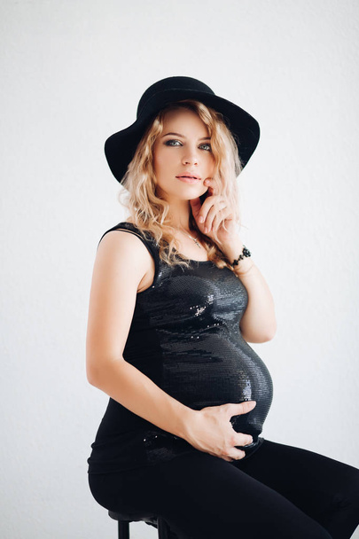 Stylish pregnant woman in black hat and sparkling top embracing her belly. - Fotografie, Obrázek