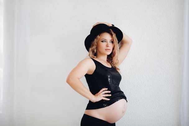 Stylish pregnant woman in black hat and sparkling top embracing her belly. - Photo, Image