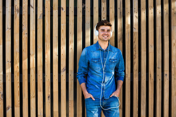 cool and modern. handsome sexy man in jeans outfit, on wooden background, with free space for text next to him, standing confident and cheerful, waiting for someone and listening music - Fotografie, Obrázek