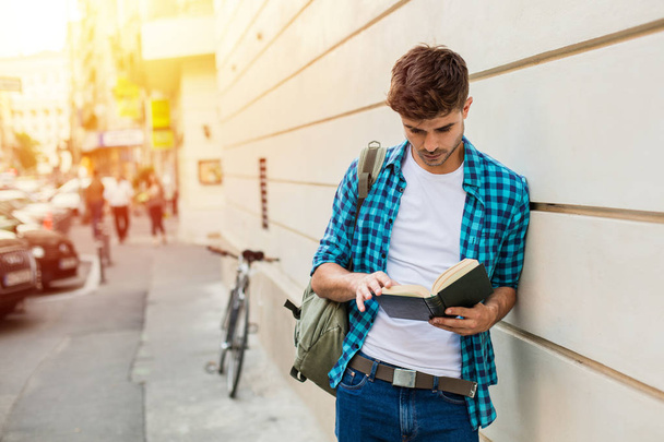 handsome young man ,student with backpack standing outside on against a wall reading a book - Photo, Image
