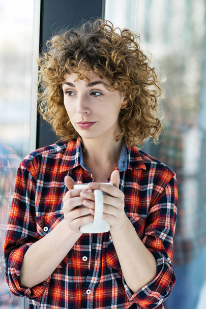 young natural curly woman casual dressed in a checkered shirt lying to a window inside his home and holding a cup of coffee or tea in hands - Photo, image