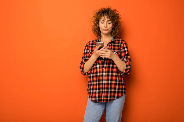 pretty curly woman in a checkered shirt and jeans standing over an orange background with empty space for text next to her with an mobile phone in hands - Fotoğraf, Görsel