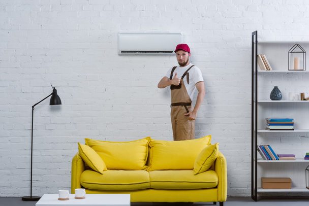 reparador profesional de pie cerca de aire acondicionado en la sala de estar y mostrando el pulgar hacia arriba
 - Foto, Imagen