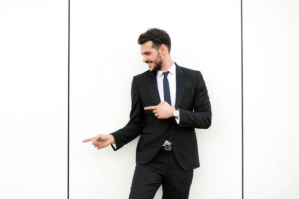 elegant man in suit, businessman, presenter, standing on white wall interacting with free space aroiund him, presenting his side with joy - Photo, Image