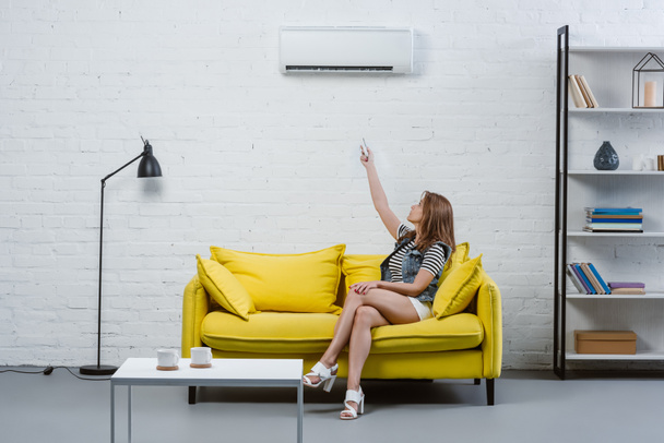 attraktive junge Frau sitzt auf Sofa und zeigt auf Klimaanlage mit Fernbedienung - Foto, Bild
