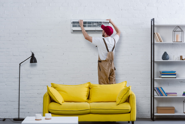 junger professioneller Reparateur repariert Klimaanlage, die an weißer Ziegelwand hängt - Foto, Bild