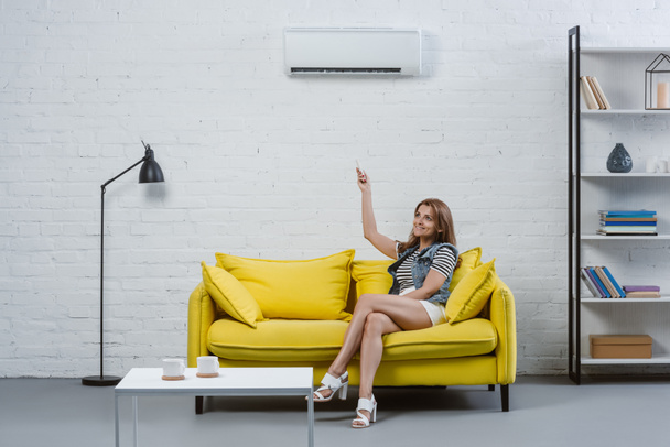 beautiful young woman sitting on sofa and pointing at air conditioner with remote control - Foto, Imagen