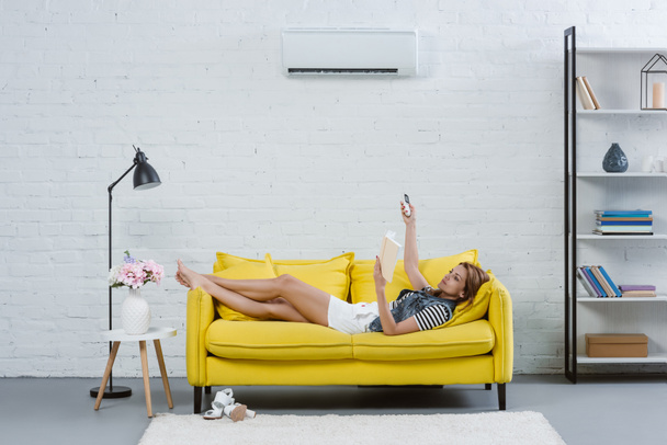 attractive young woman reading book on sofa and pointing at air conditioner with remote control - Photo, Image